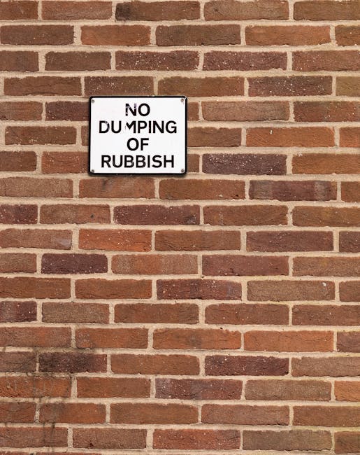 A close-up view of a rectangular white metal sign mounted on a red brick wall. The sign displays the message 'NO DUMPING OF RUBBISH' in black capital letters, with some letters slightly faded or worn.