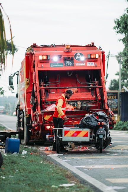A worker wearing a high-visibility yellow and orange vest operates a red rubbish collection truck, positioned on the roadside during daytime. The rear of the truck, which features a large hydraulic co