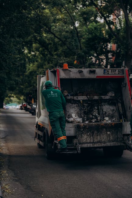 A worker wearing a high-visibility yellow and orange vest operates a red rubbish collection truck, positioned on the roadside during daytime. The rear of the truck, which features a large hydraulic co